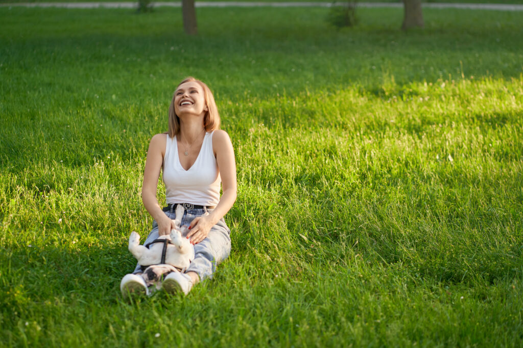 Young laughing woman sitting on grass with male french bulldog on legs, having fun. Front view of gorgeous caucasian caucasian girl enjoying summer warm day with dog, petting his belly in city park.
